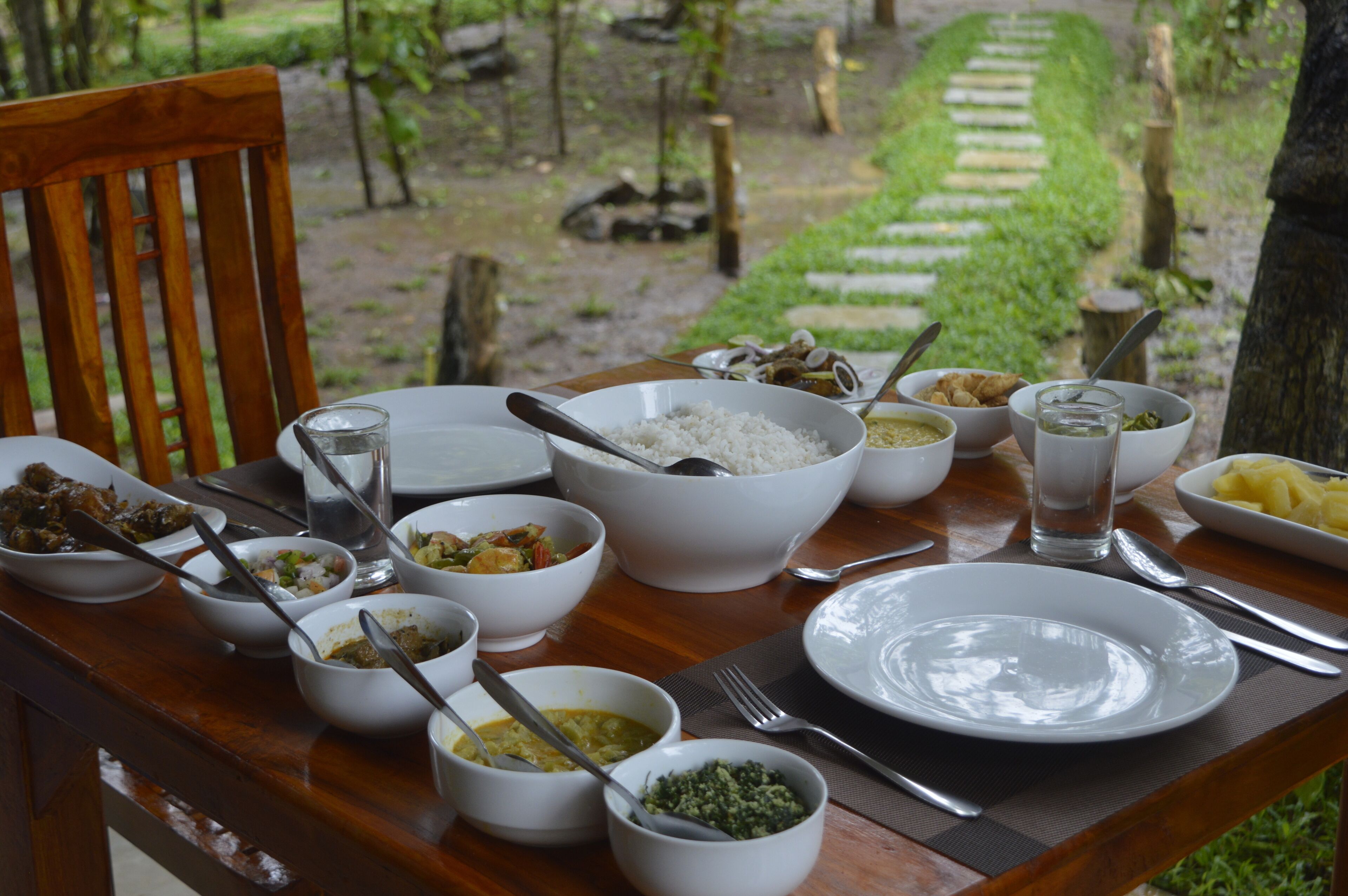 Une table à manger dressée avec différents plats et couverts, entourée d’un cadre naturel en plein air.
