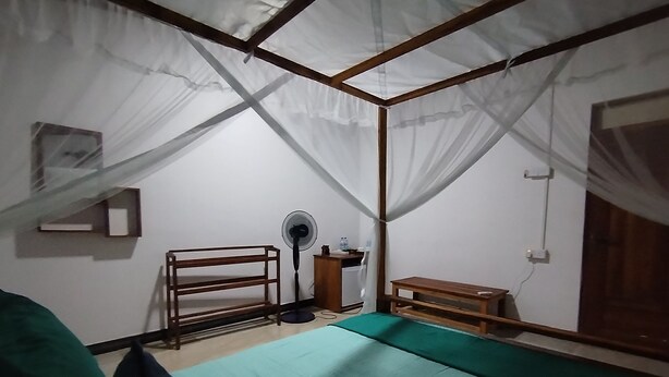 Una habitación con cama, escritorio, silla, ventilador de pie y mosquitero.