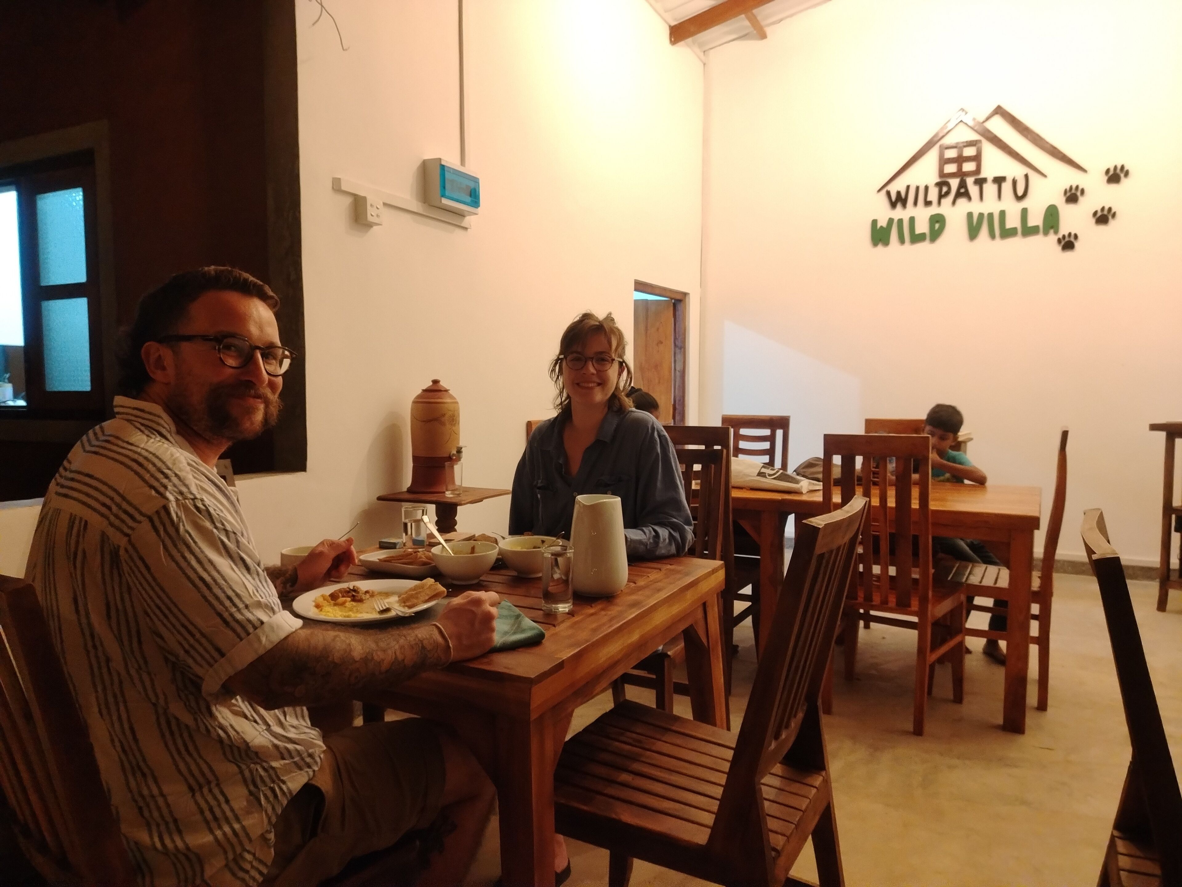 A dining area with wooden tables and chairs, a wall-mounted thermometer, and a logo for 'Wilpattu Wild Villa'.