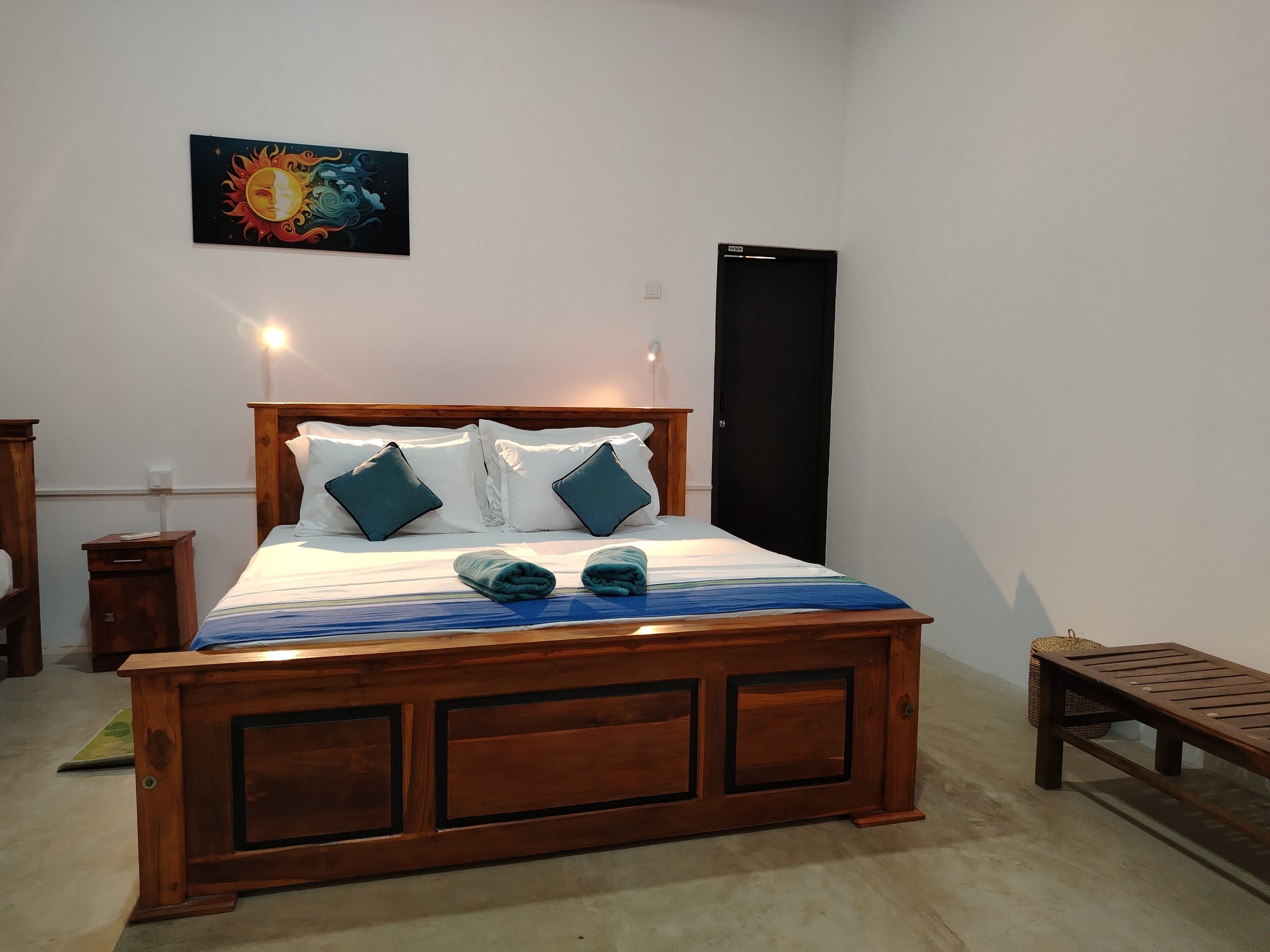 A bedroom with a wooden bed, two pillows, a bedside table, a nightstand, a wall-mounted artwork, a door, and a bench.
