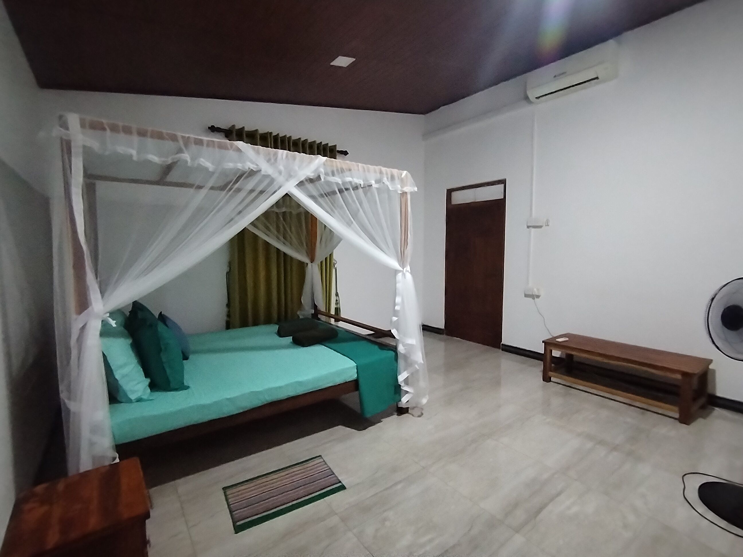 Una stanza con un letto a baldacchino, una panca, una ventola e una porta.