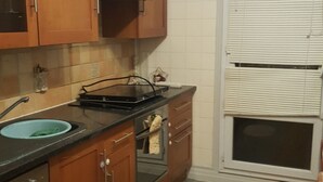 Une cuisine de taille réduite, dotée de meubles en bois, d’un plan de travail noir, d’un évier et d’une fenêtre avec des stores.