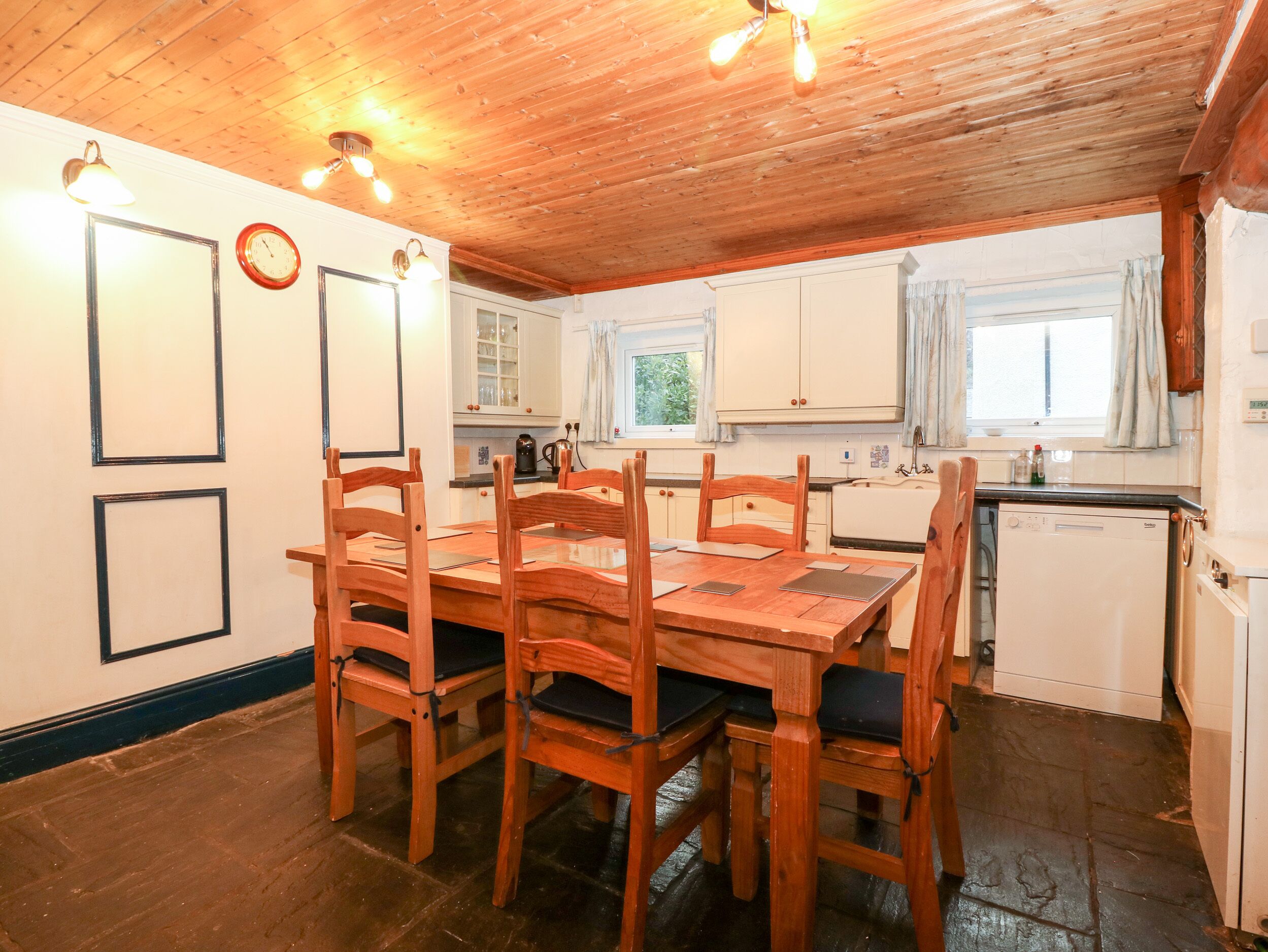 Une table à manger en bois avec des chaises, une cuisine avec des meubles blancs, une horloge et une fenêtre avec des rideaux.
