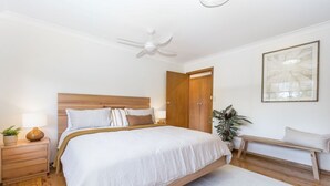 A bedroom with a large bed, a ceiling fan, a wooden headboard, a bedside table with a lamp, a bench, and a potted plant. - Byron's Beach House Mylestom
Entire home in Mylestom, Australia (Mylestom)