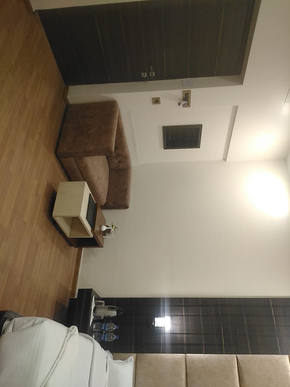 A hotel room with a brown sofa, a small table with a lamp, a bed, and a dark tiled wall.