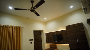 A hotel room with a ceiling fan, a flat-screen TV mounted on a wooden panel wall, a brown armchair, and a small table with a remote control. - Hotel New Grand (Deoghar)