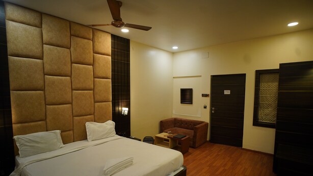A hotel room with a large bed, a ceiling fan, a small seating area, and a door with a window. - Hotel New Grand (Deoghar)
