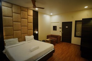 A hotel room with a large bed, a ceiling fan, a small seating area, and a door with a window. - Hotel New Grand (Deoghar)