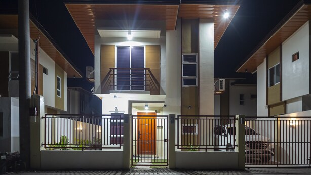 A modern two-story building with a central entrance, a balcony on the upper floor, and a gated driveway.