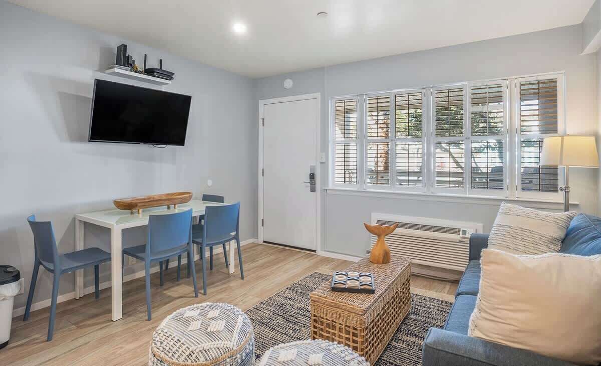 A modern living room with a flat-screen TV, a dining area with chairs, a sofa, and a large window with blinds.