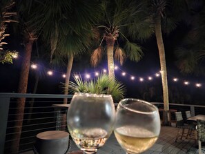 A deck with two wine glasses, palm trees, and string lights. - 116 Sea Otter - Resort Access & Golf Cart! (Fripp Island)