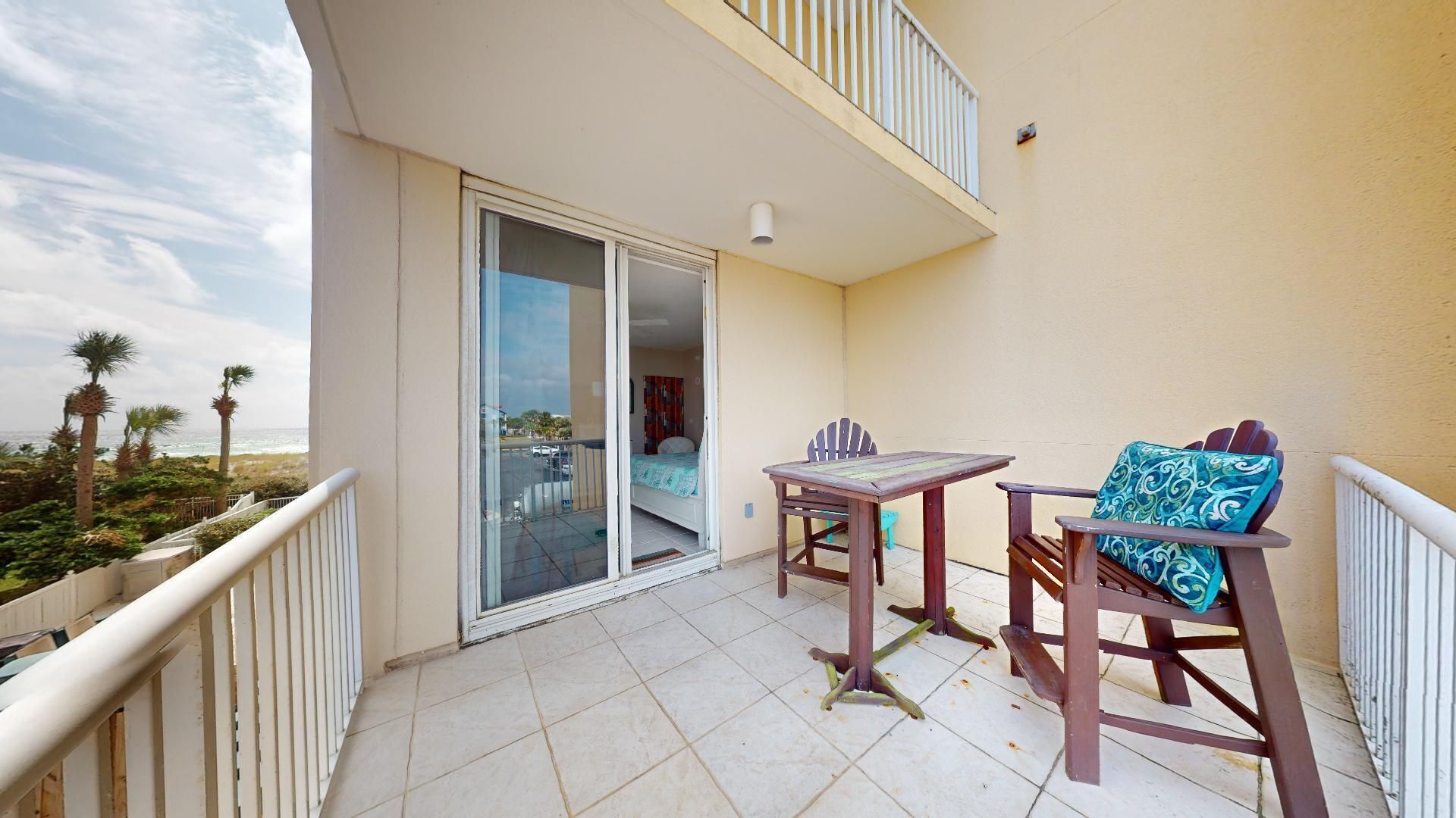 A balcony with a table and chairs, a sliding glass door leading to a bedroom, and a view of the beach.