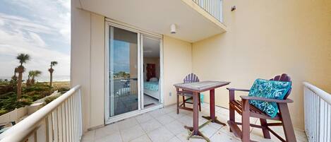 A balcony with a table and chairs, a sliding glass door leading to a bedroom, and a view of the beach.