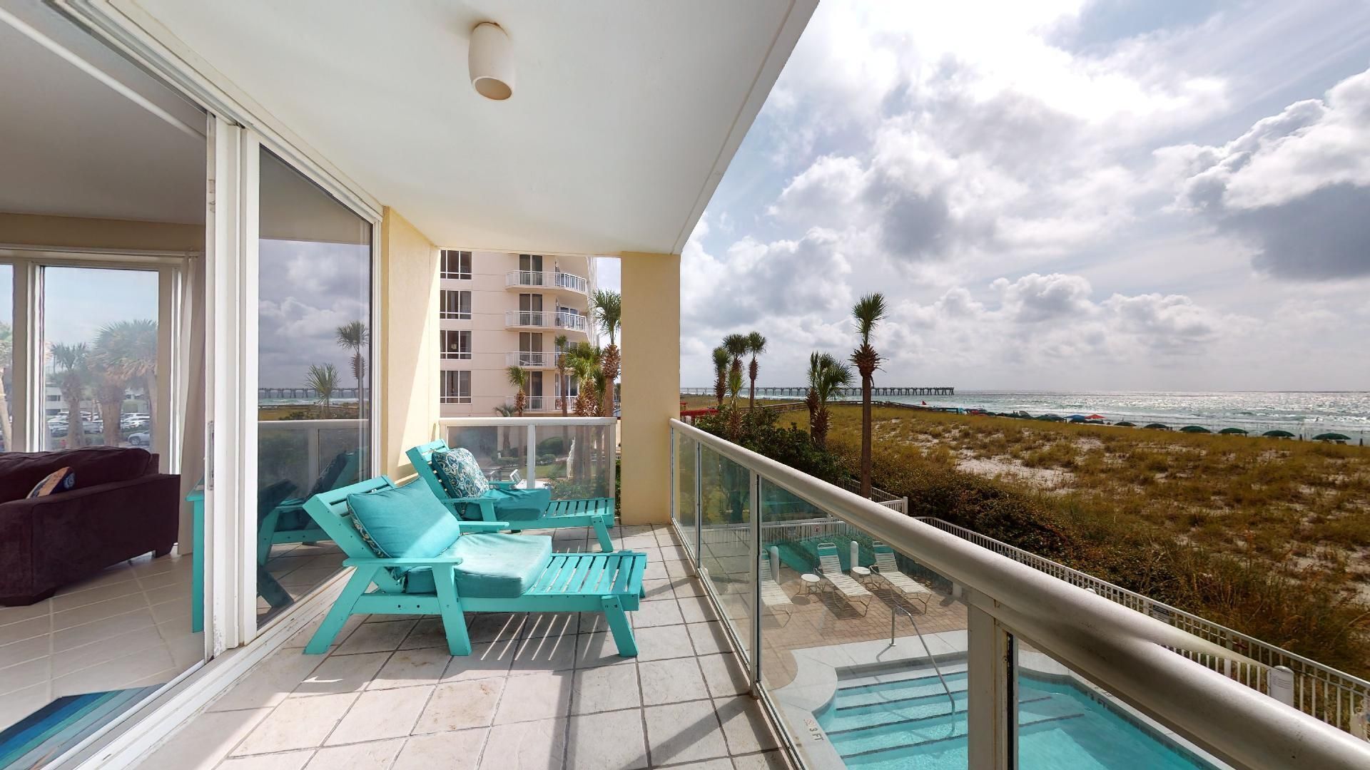 Ein Balkon mit türkisfarbenen Liegestühlen, einer Glasreling und Blick auf Strand und Meer.
