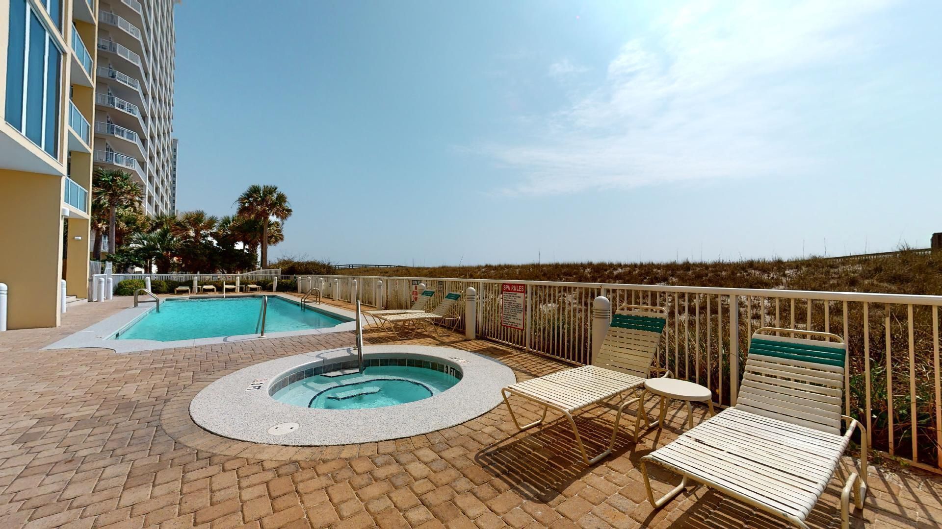 A rooftop pool area with a hot tub and lounge chairs, surrounded by a fence and palm trees.