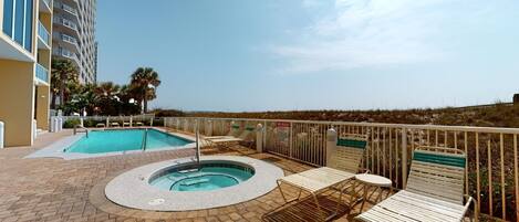A rooftop pool area with a hot tub and lounge chairs, surrounded by a fence and palm trees.