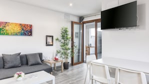 Una sala moderna con un sofá gris, una mesa de centro blanca, una mesa de comedor con sillas, una planta en maceta y un televisor de pantalla plana montado en la pared.