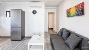 Un espacio de estar moderno con un sofá gris, una mesa de centro blanca, un refrigerador de acero inoxidable, un cuadro abstracto colorido y un reloj en la pared.