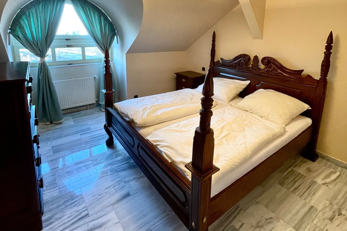 A bedroom with a wooden four-poster bed, a radiator, a dresser, and a window with curtains.