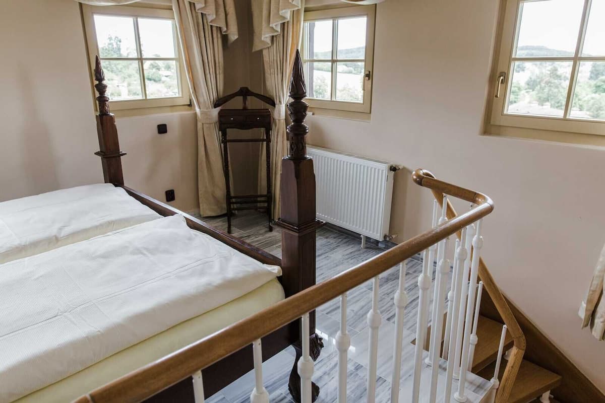 A bedroom with a wooden four-poster bed, a small bedside table, a radiator, and a window with curtains.