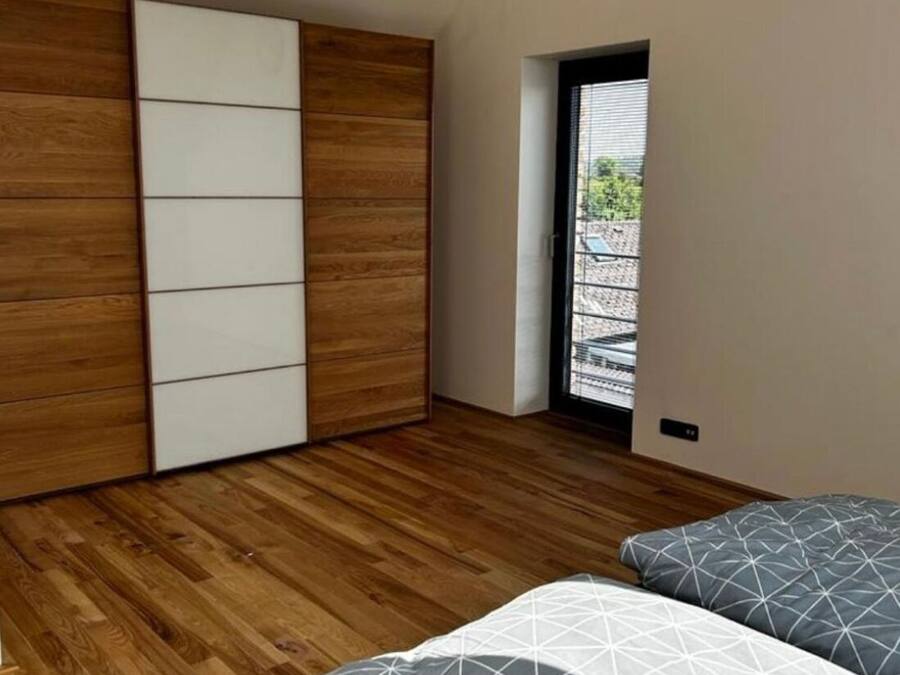 A bedroom with a wooden floor, a bed with patterned bedding, a wardrobe with sliding doors, and a view through a door to the outside.