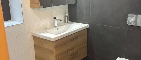 Un baño moderno con lavamanos blanco, tocador de madera, paredes de azulejos oscuros y un inodoro blanco.