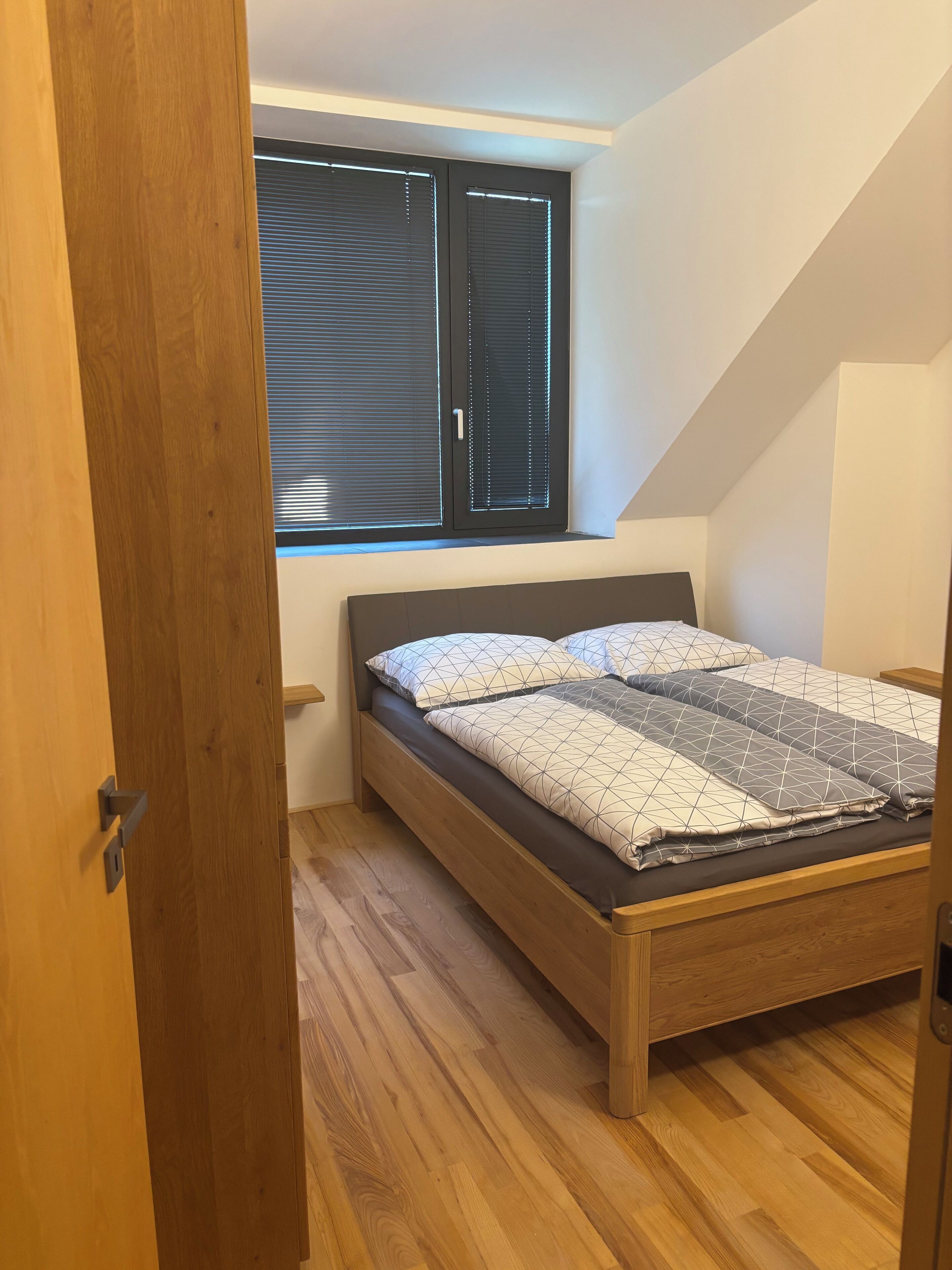 Een slaapkamer met een houten bed, een raam met zonwering en een schuine kap.