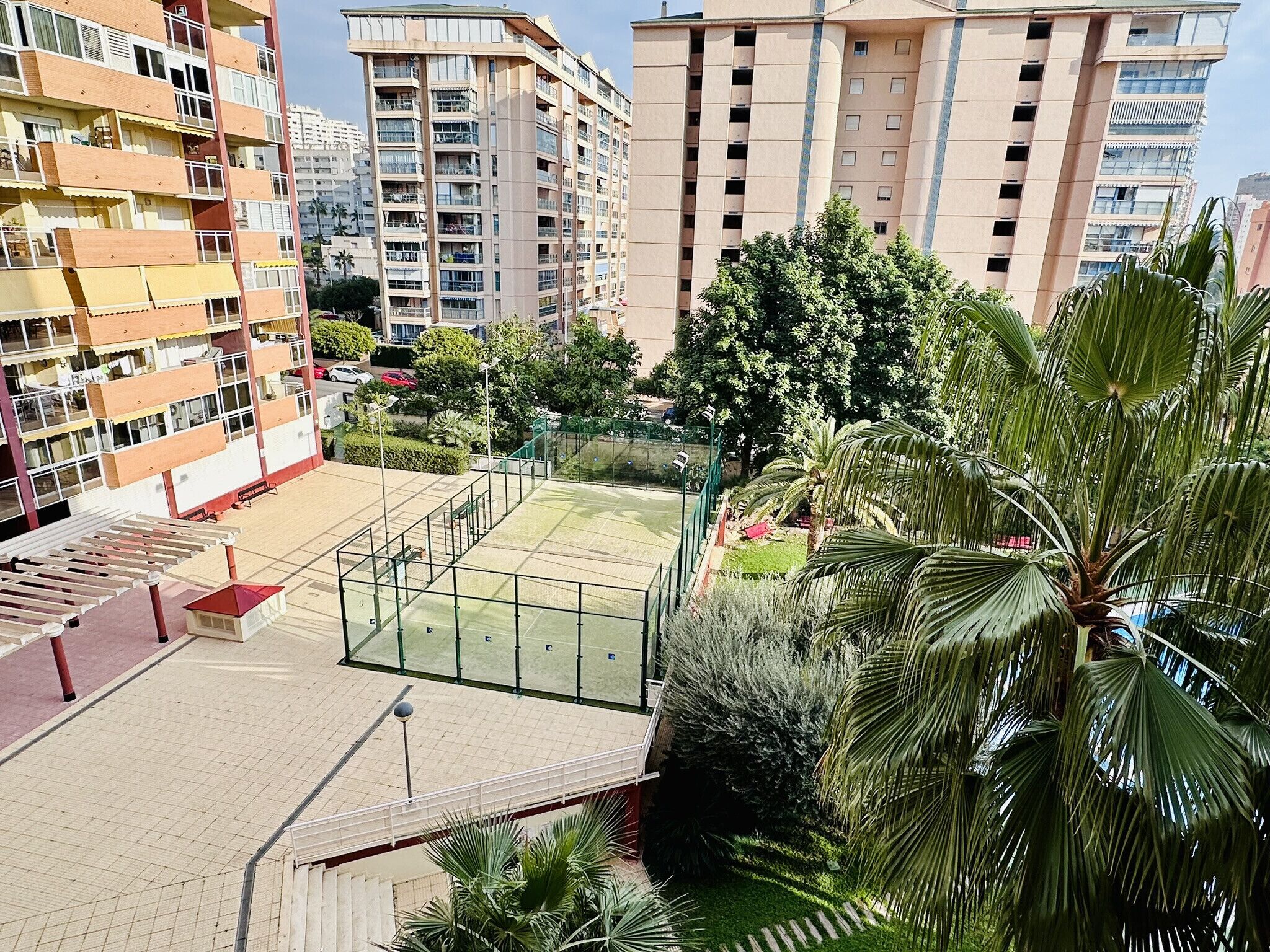 A multi-story residential building with balconies, a courtyard with a tennis court, and surrounding greenery.