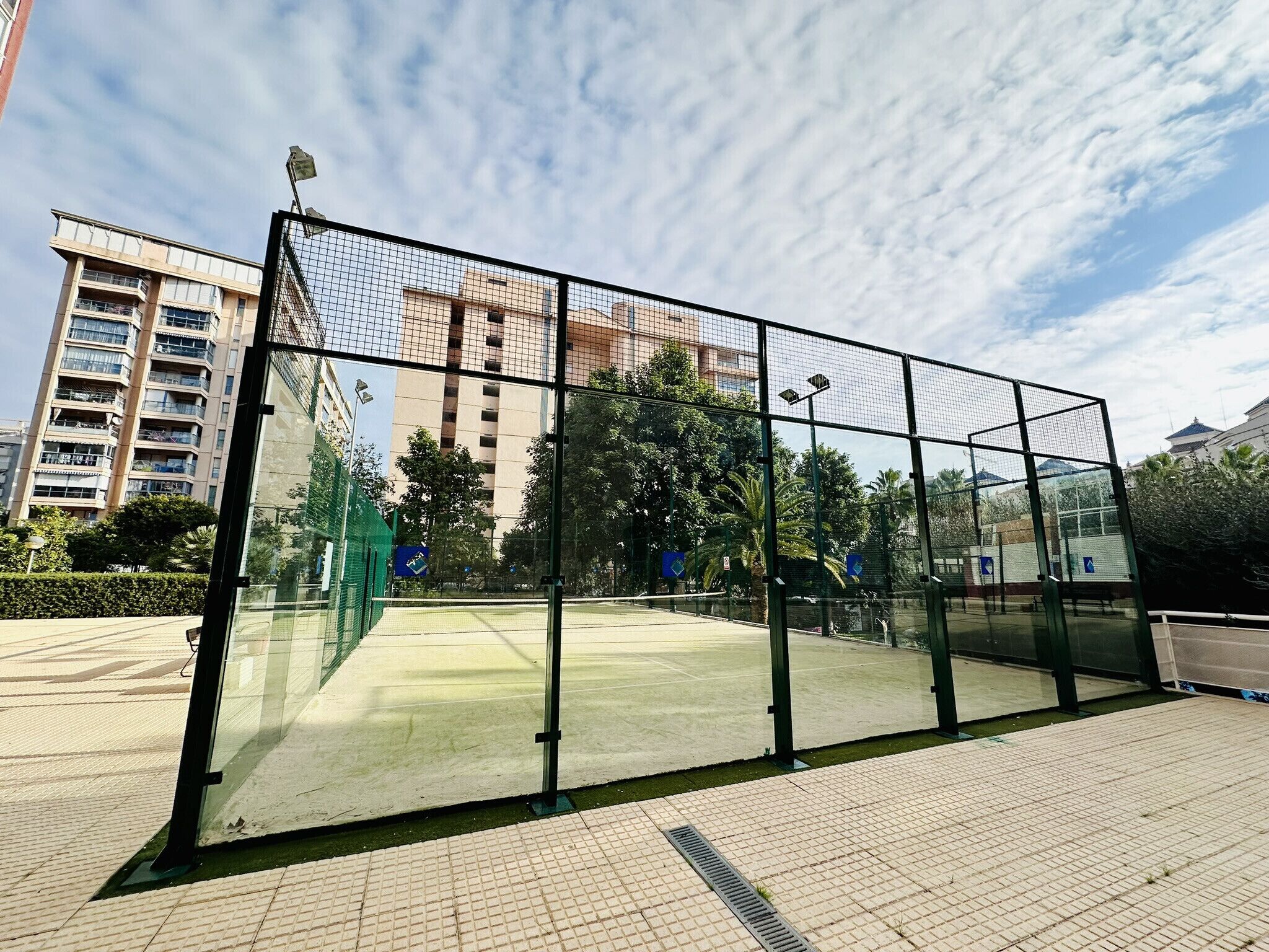 A sports court with a glass enclosure, surrounded by a paved walkway and greenery.