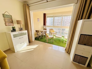 A room with a dining table, chairs, a small fan, a white cabinet, and a window with a view of buildings.
