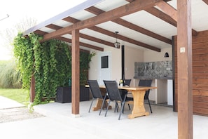 Un patio exterior con mesa y sillas de madera, una zona de cocina con salpicadero de azulejos y una estructura de pérgola de madera.