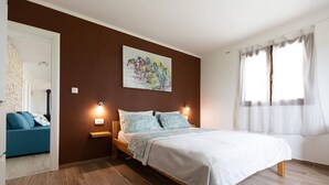 Ein Schlafzimmer mit Bett, Nachttisch, Wandbild, Fenster mit Vorhängen und einem blauen Sofa im angrenzenden Raum.