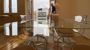 A modern dining area with a glass table and metal chairs, a vase of white flowers, and a view of buildings through a doorway.