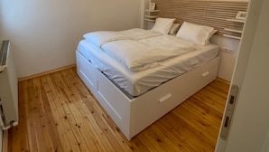 A bedroom with a wooden floor, a white bed with a wooden headboard, and a ceiling light fixture.