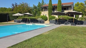 Ein Pool mit Liegestühlen und Sonnenschirmen, eine gepflegte Rasenfläche und im Hintergrund ein Gebäude mit einem Ziegeldach.