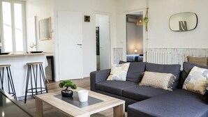 A modern living room with a grey sofa, a wooden coffee table, and a small potted plant. There is a kitchen area with stools and a large mirror on the wall. - Villa Gaby ~ Rare 6 Pers ~ Hypercentre (Pau)