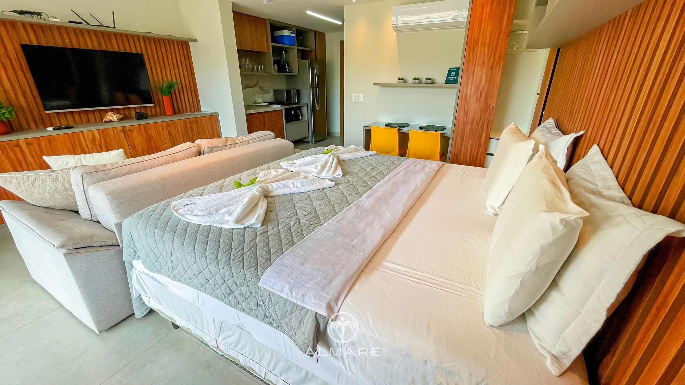 A modern hotel room with a large bed, a flat-screen TV mounted on the wall, a kitchenette, and a bathroom visible through an open door.