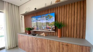 A modern living room with a wooden cabinet, a flat-screen TV mounted on a wooden wall, a potted plant, and a decorative shell. - Flat Ilê de Pipa Oásis - By Almare (Tibau do Sul)