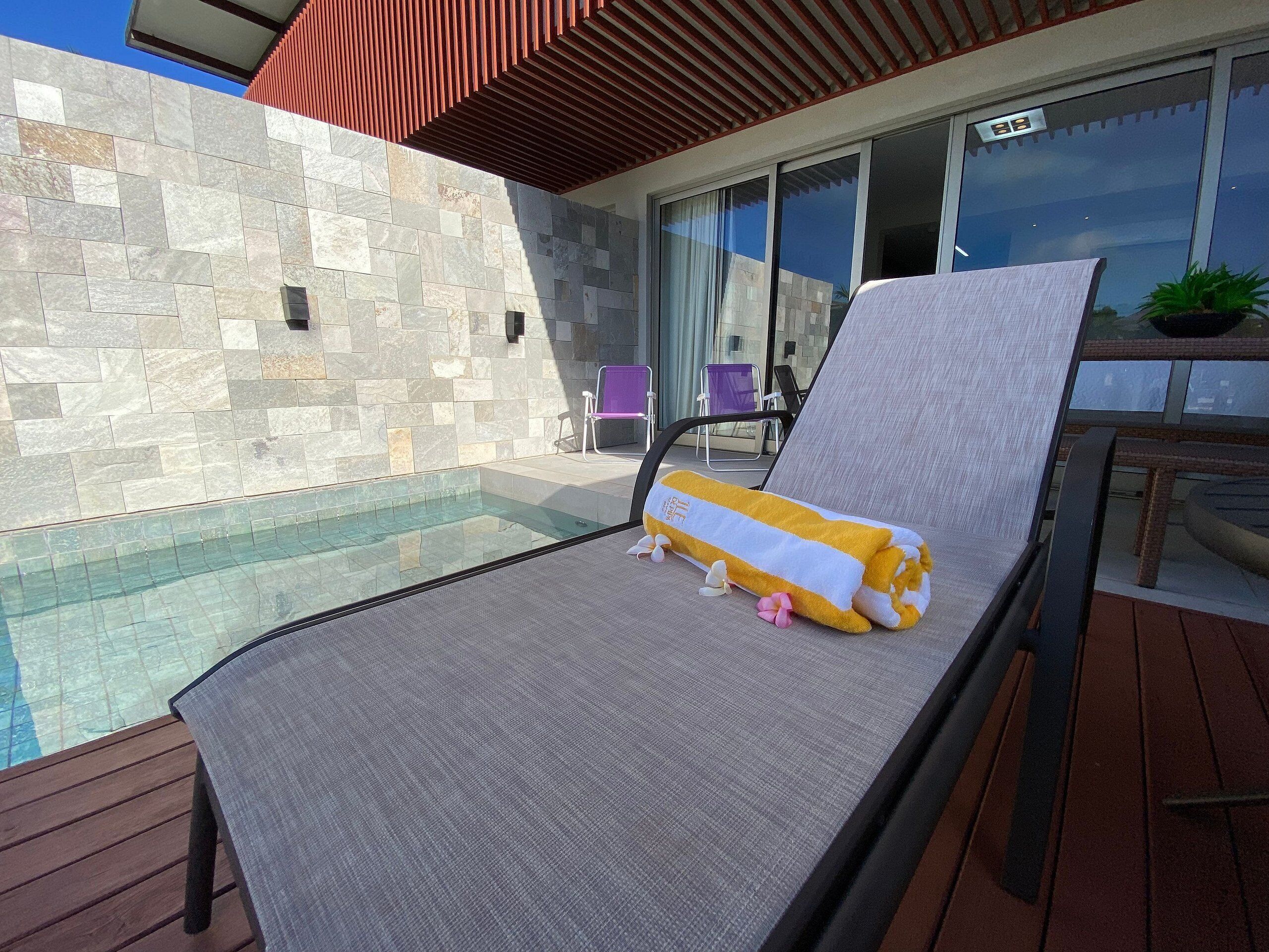 A poolside lounge chair with a towel and plush toy, a stone wall, and a glass door leading to a patio area.