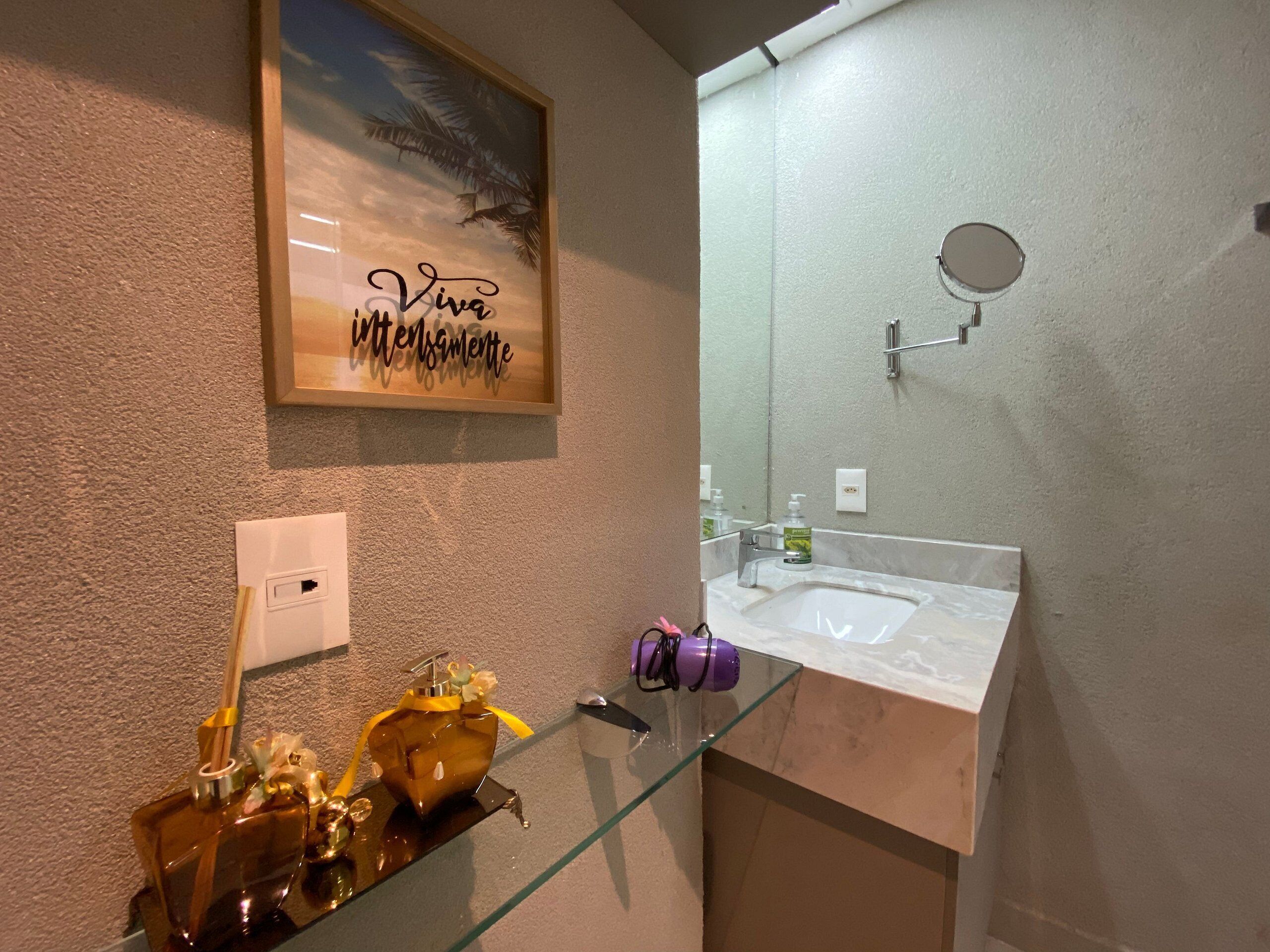 Une salle de bain avec une étagère en verre où sont posés un flacon de parfum et un sèche-cheveux, un lavabo avec un miroir au-dessus, et un tableau encadré accroché au mur.