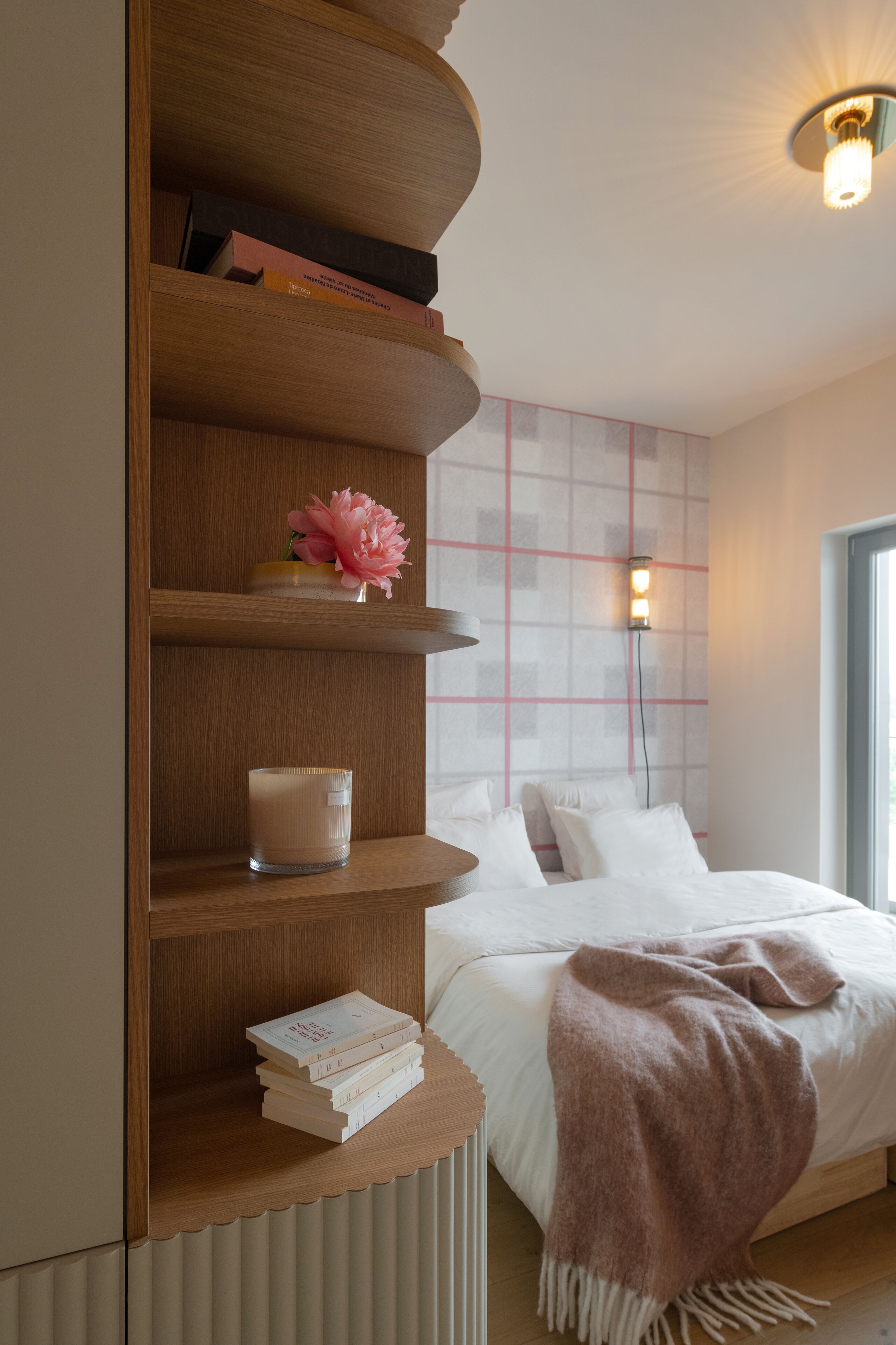 Een slaapkamer met een bed, een nachtkastje met boeken en een kaars, een wandlamp en een raam.