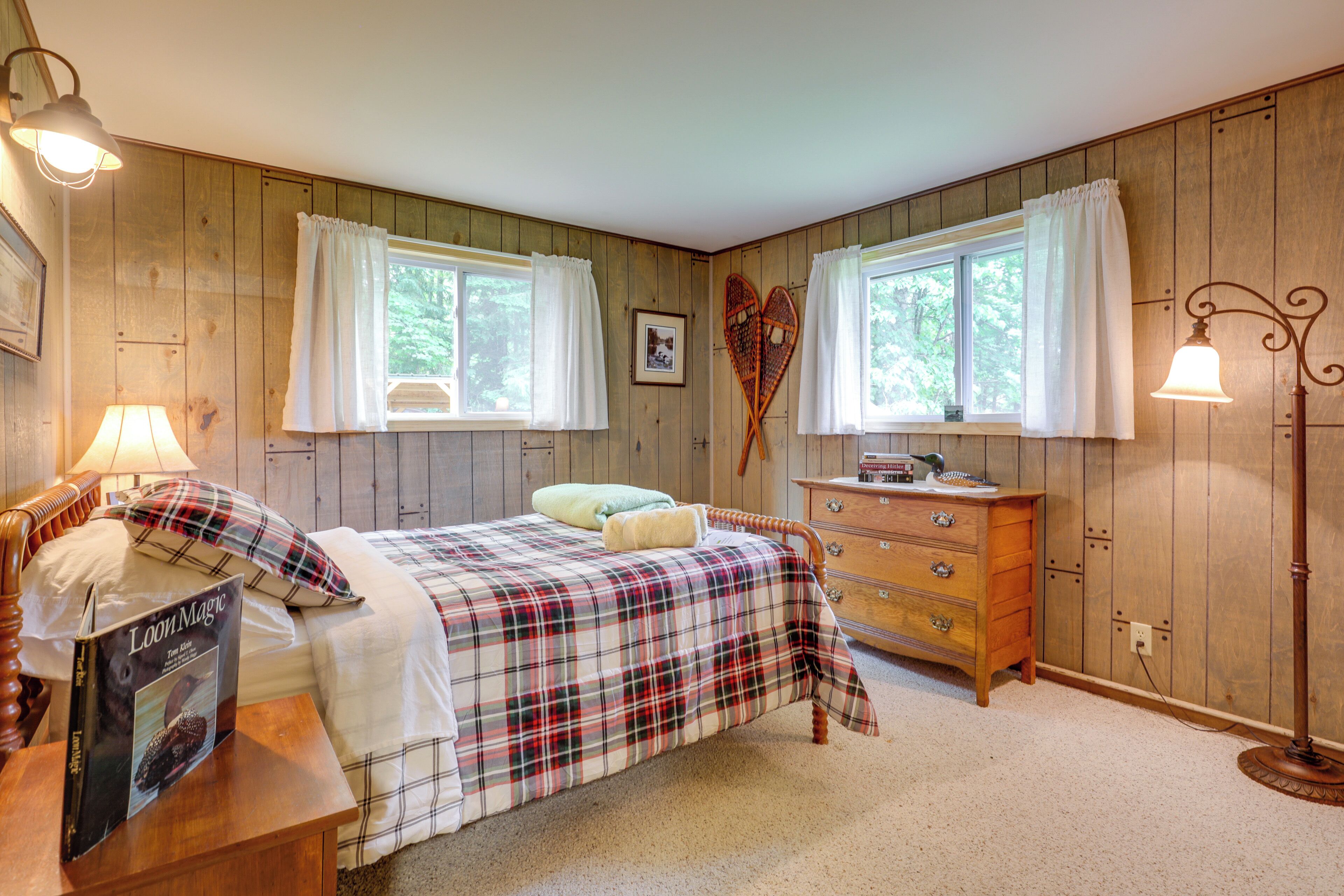 A bedroom with a plaid bedspread, wooden walls, two windows with white curtains, a dresser, and a bedside lamp.