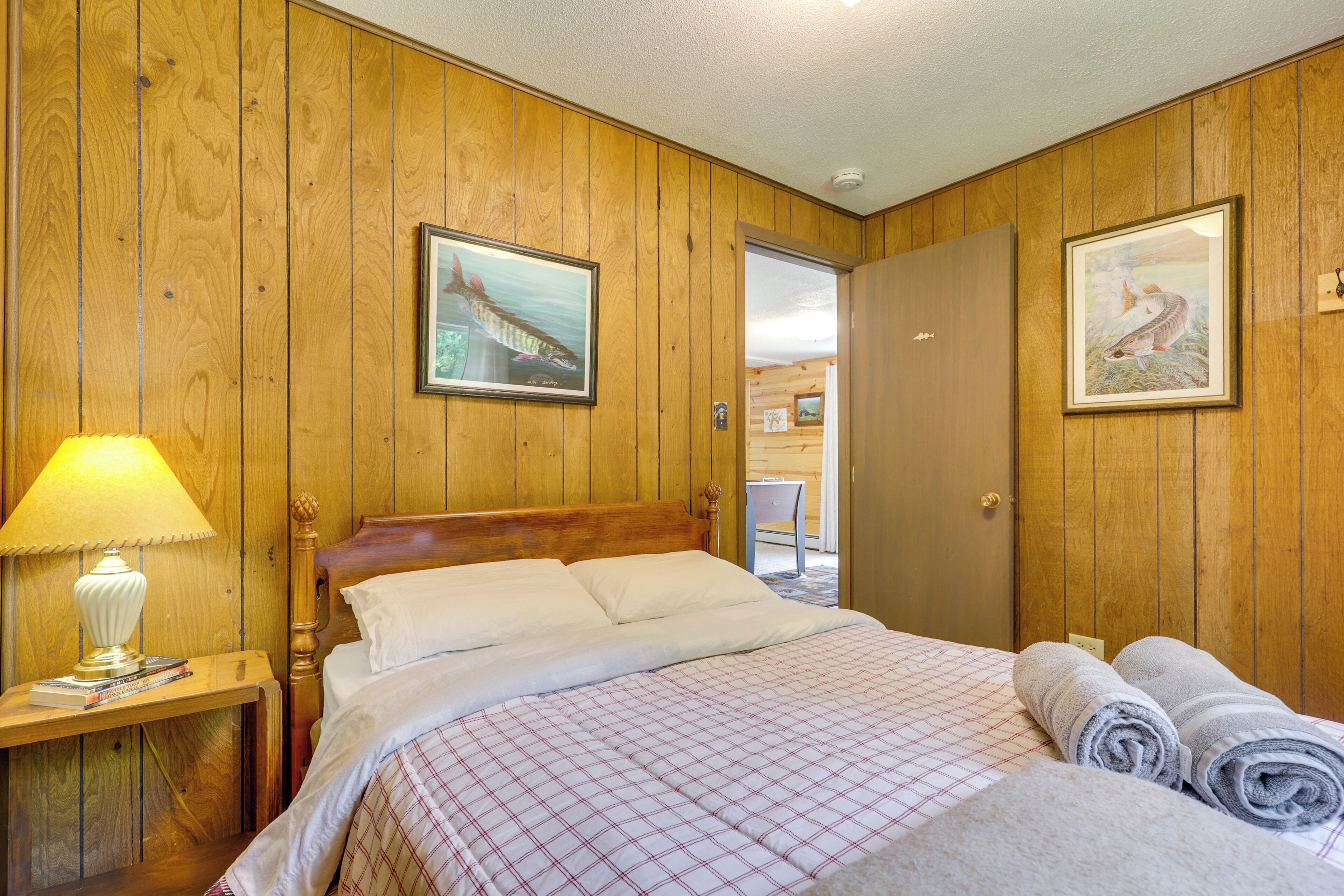 Een kamer met houten lambrisering, een bed, een nachtkastje met een lamp en twee ingelijste foto’s aan de wanden.