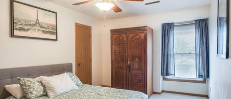 Un dormitorio con cama, ventilador de techo, armario y ventana con cortinas.