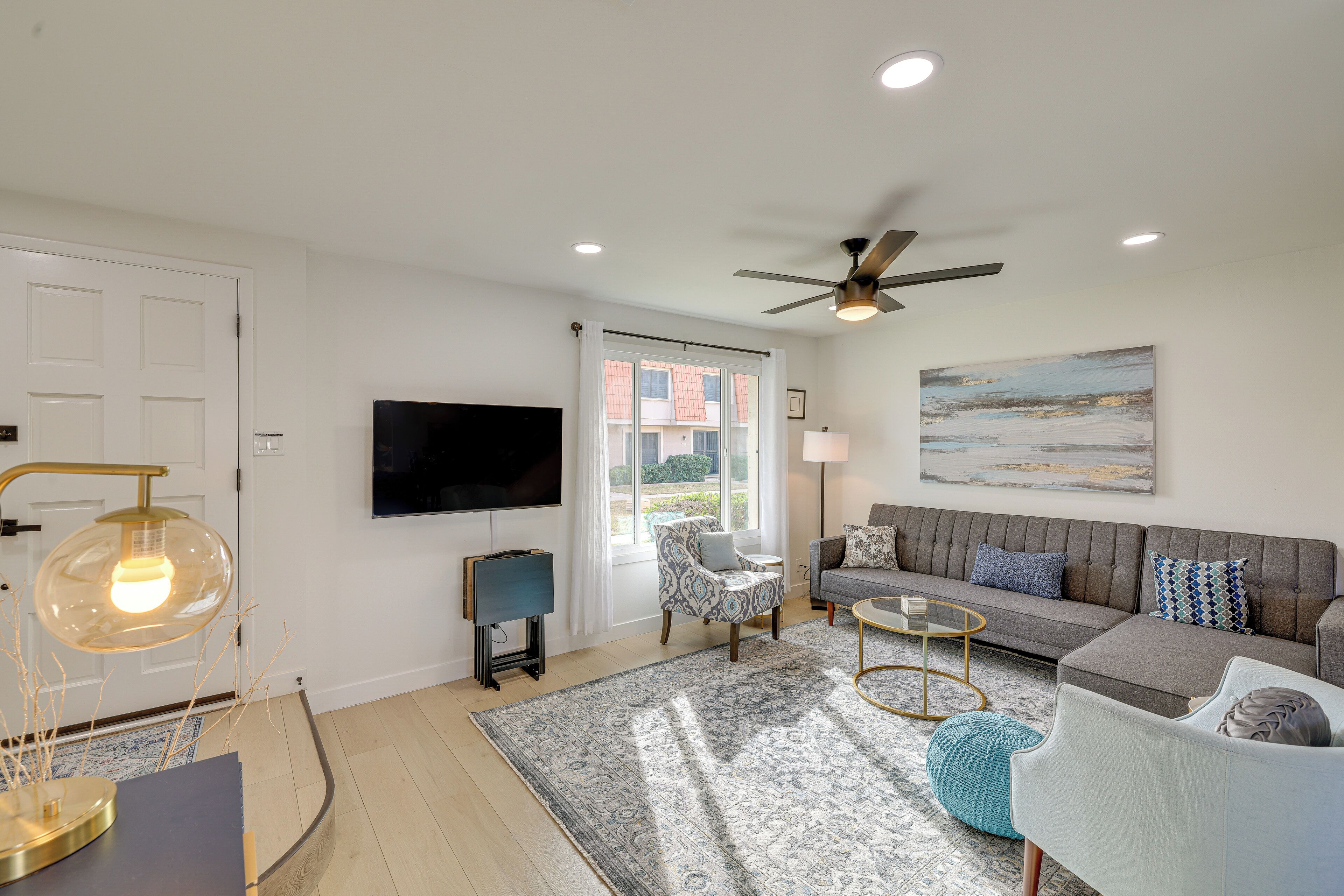 A modern living room with a grey sofa, patterned armchair, and a blue armchair. There is a flat-screen TV mounted on the wall, a ceiling fan, and a large window with curtains.