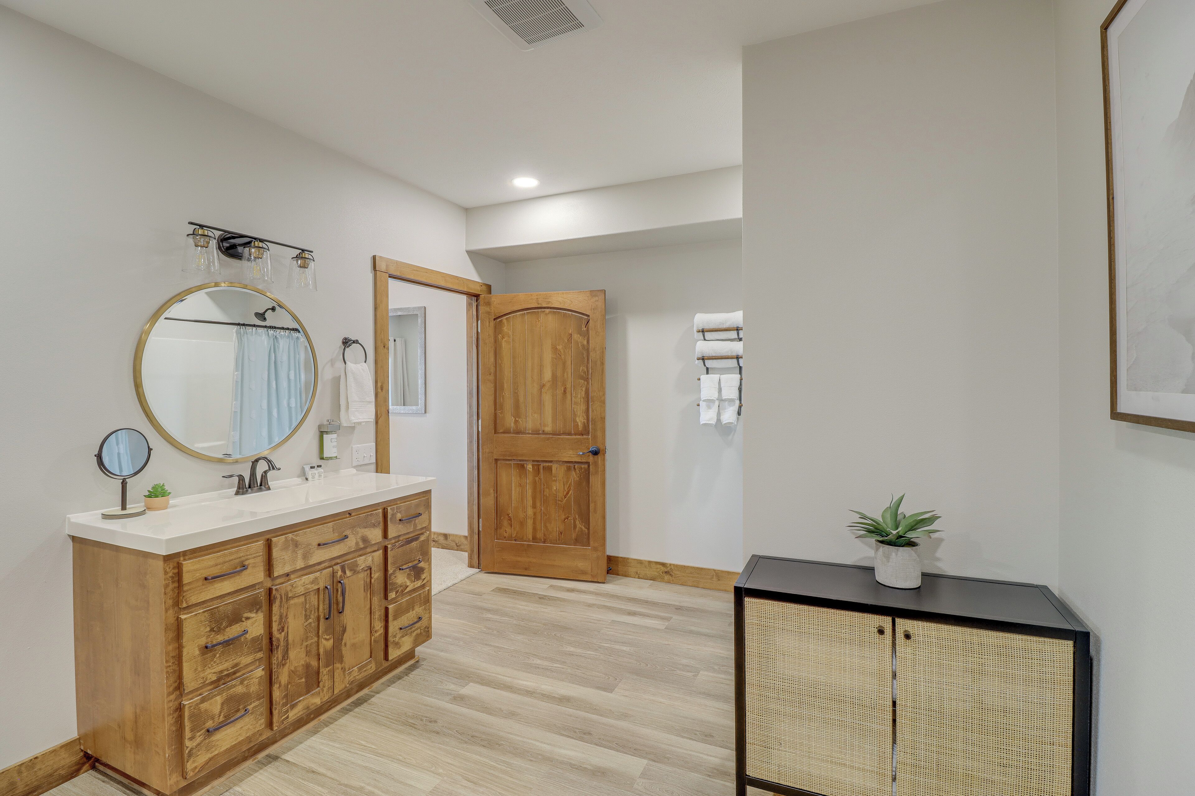 A bathroom with a wooden vanity, a round mirror, a shower curtain, and a potted plant on a black console.