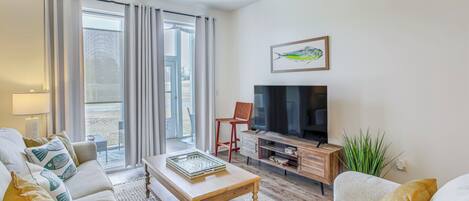 Una sala de estar moderna con sofá, mesa de centro, televisor y un amplio ventanal con cortinas.