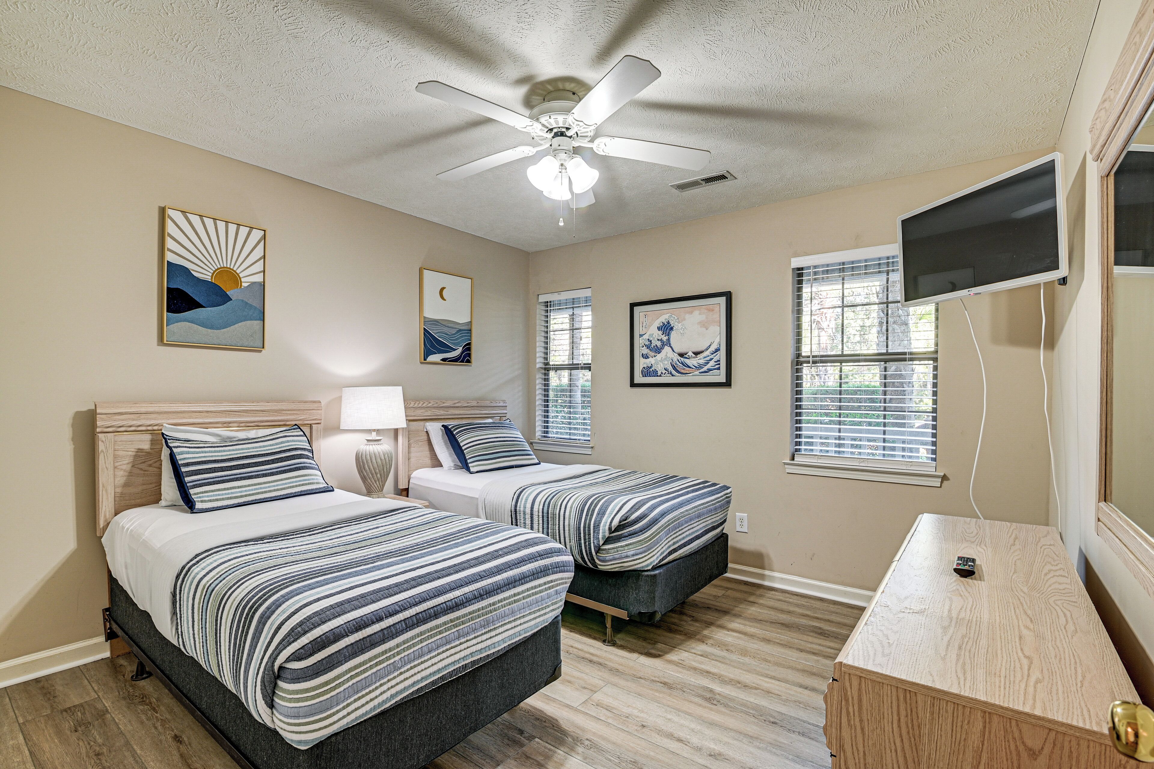 A bedroom with two beds, a ceiling fan, artwork on the walls, a TV, and a wooden dresser.