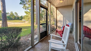 A covered porch with a view of a golf course, a white bench, and a red chair. - Pawleys Island Condo w/ Screened Porch + Golfing! (Pawleys Island)
