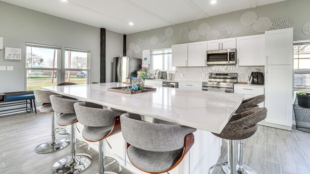 A modern kitchen with a large island, bar stools, stainless steel appliances, and decorative wall art. - Modern Farmhouse in Del Valle: Rural Setting! (Del Valle)